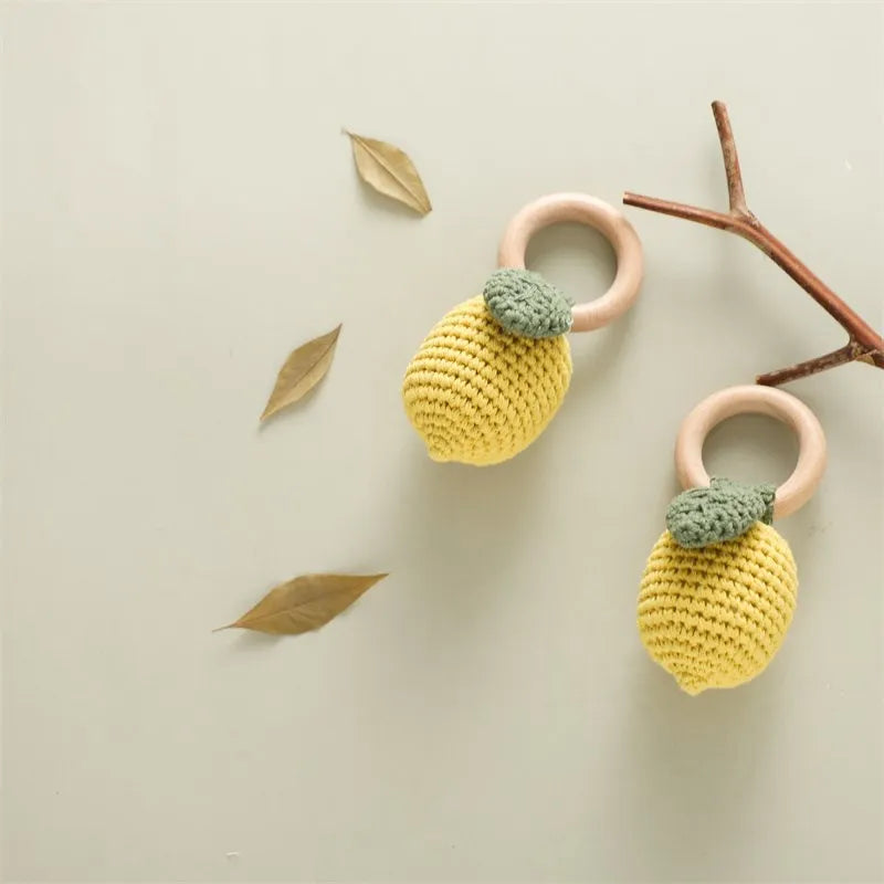Two yellow crochet lemon toys with green leaves on pink rings, placed on a light beige surface with leaves and a branch.