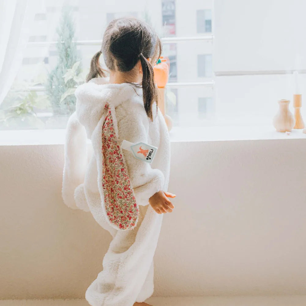 Child wearing a fluffy white onesie with floral patterned bunny ears in a bright room.